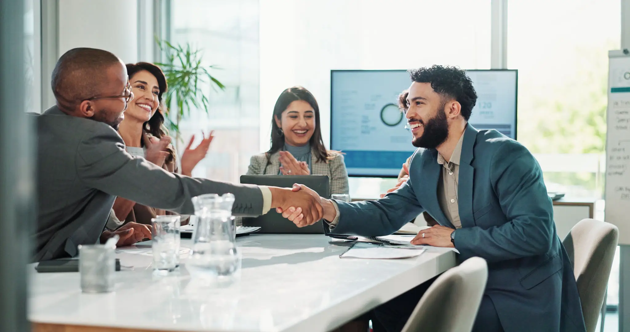 Two people shaking hands across a meeting table with colleagues smiling.